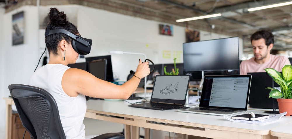 Eine junge Frau sitzt vor einem Laptop. Sie trägt eine Virtual Reality Brille und gestaltet mit Gesten einen Schuh, der auf dem Bildschirm zu sehen ist.
