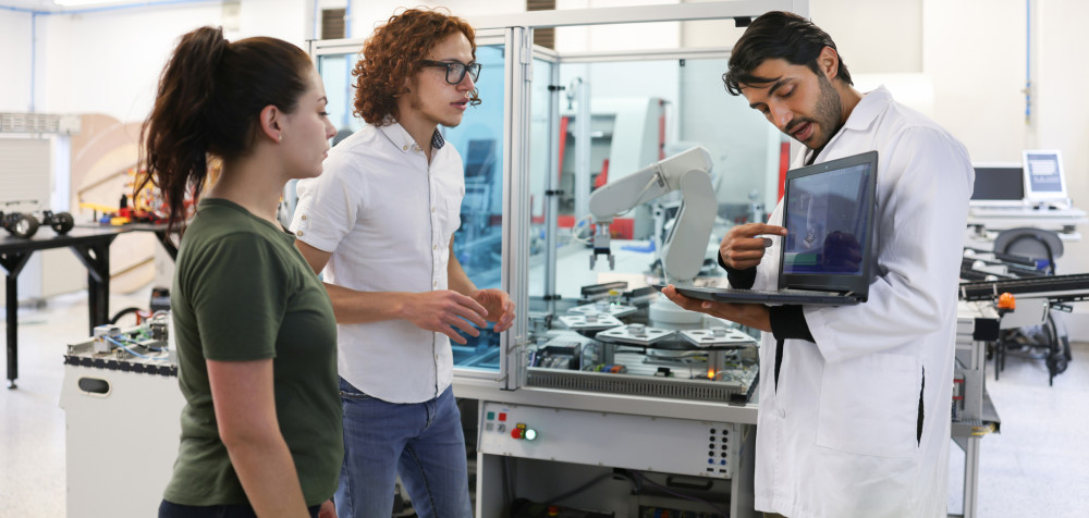 Ein junger Mann zeigt auf einem Laptop einer jungen Frau und einem jungen Mann etwas. Sie stehen inmitten eines Labors mit modernen Maschinen.