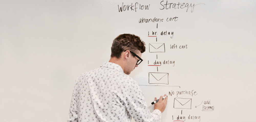 Ein junger Mann steht vor einem Whiteboard und schreibt die Schritte einer Workflow Strategy an.