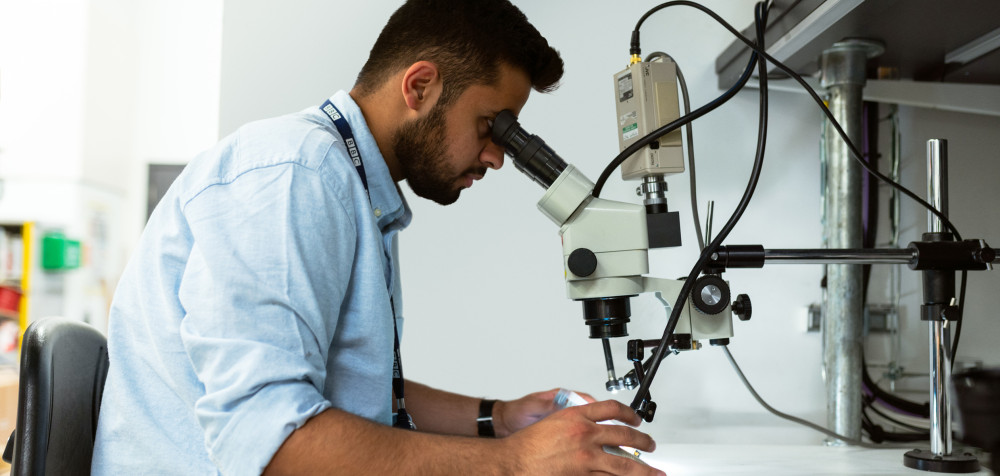 Ein junger Mann sitzt an einem Mikroskop und schaut sich eine Probe an.