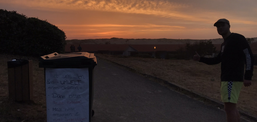 Junger Mann bei Sonnenuntergang. Beschriftete Mülltonne daneben.