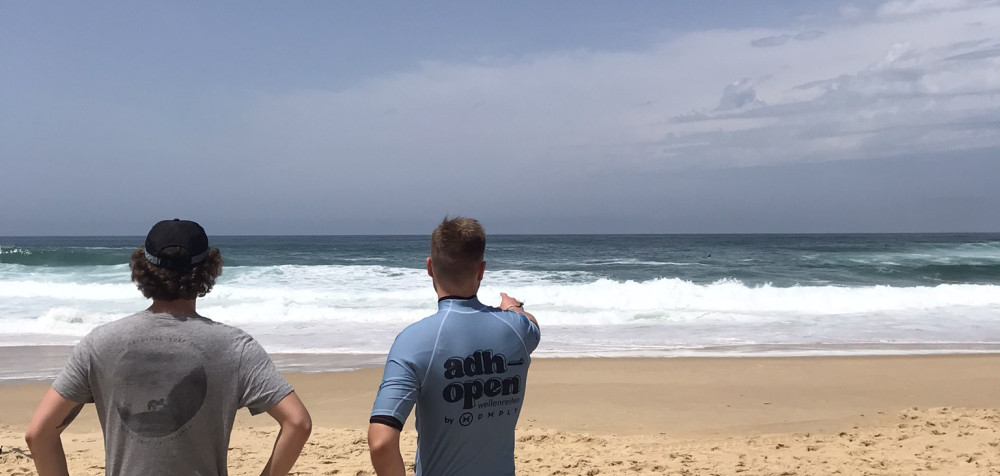 zwei junge Surfer stehen mit dem Rücken zur Kamera am Strand und blicken zum Meer